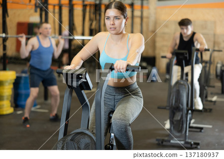 Motivated young woman practicing on exercise bike near other people in gym Motivated young woman practicing on exercise bike near other people in gym 137180937