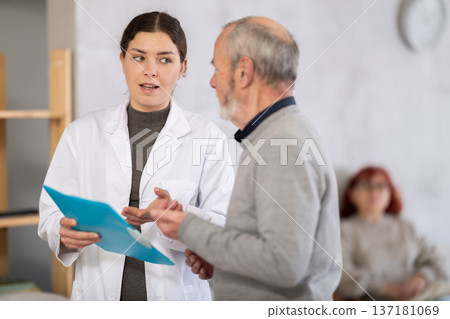 Positive polite nurse or doctor talking to aged man patient in reception area in background queue of people 137181069