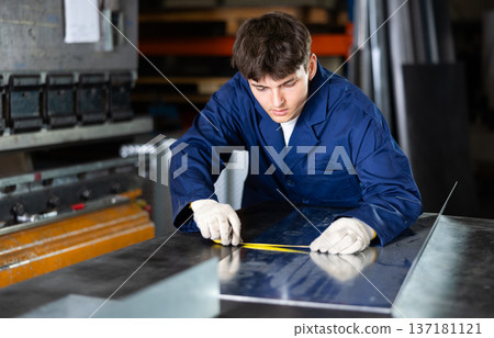 Guy working at metallurgical plant measures sheet of iron using tape measure and marks cutting line Guy working at metallurgical plant measures sheet of iron using tape measure and marks cutting line 137181121