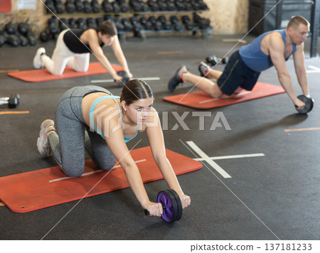 Motivated young woman practicing exercises with abs roller near other people in gym 137181233