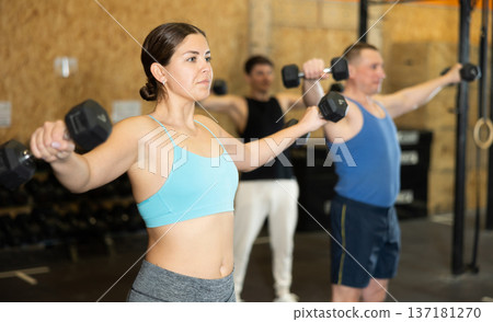 Motivated young woman practicing exercises with dumbbells near other people in gym Motivated young woman practicing exercises with dumbbells near other people in gym 137181270