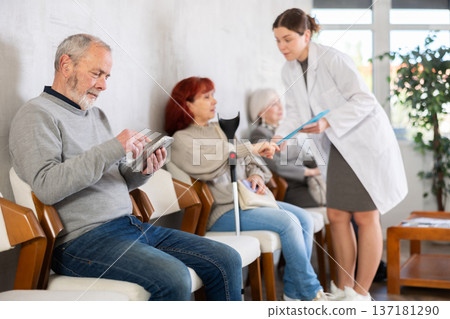 Elderly man sitting in clinic queue while nurse talks to other patients 137181290