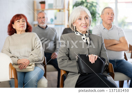 Cheerful and sociable mature woman sitting on chairs, listening lecture and laughing with group of students of different ages in lecture hall Cheerful and sociable mature woman sitting on chairs, listening lecture and laughing with group of students of different ages in lecture hall 137181450