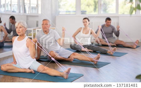 Elderly woman performs stretches elastic band with hand and foot while sitting on mat. 137181481