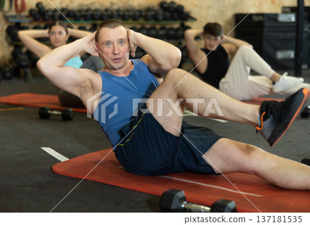 Dedicated man practicing knee raises near other people in gym Dedicated man practicing knee raises near other people in gym 137181535