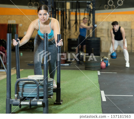 Concentrated young girl performing intense sled pushing in gym Concentrated young girl performing intense sled pushing in gym 137181563