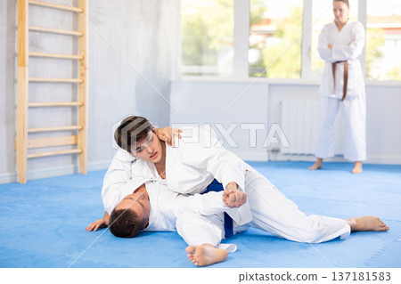 Judo or jiu-jitsu classes - two men practicing grabbing and throwing on sports mats under the guidance of trainer Judo or jiu-jitsu classes - two men practicing grabbing and throwing on sports mats under the guidance of trainer 137181583