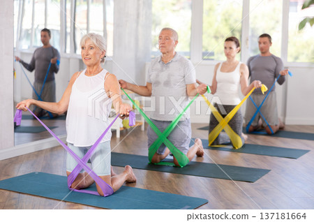Group of seniors using resistance bands to stretch their arms and body while kneeling on mats in gym Group of seniors using resistance bands to stretch their arms and body while kneeling on mats in gym 137181664
