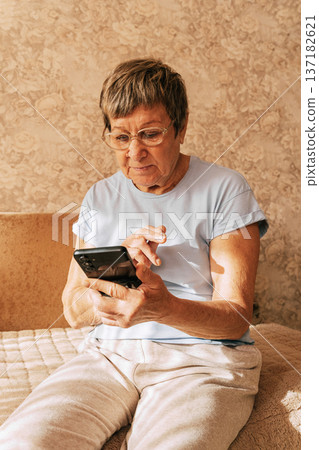 An elderly woman wearing glasses was intently immersed in her smartphone while sitting on a sofa in a sun-drenched. Digital Phone Addiction in Older Women, Screen Time Overload, and Modern Technology An elderly woman wearing glasses was intently immersed in her smartphone while sitting on a sofa in a sun-drenched. Digital Phone Addiction in Older Women, Screen Time Overload, and Modern Technology 137182621
