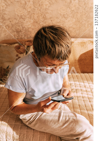 An elderly woman wearing glasses was intently immersed in her smartphone while sitting on a sofa in a sun-drenched. Digital Phone Addiction in Older Women, Screen Time Overload, and Modern Technology An elderly woman wearing glasses was intently immersed in her smartphone while sitting on a sofa in a sun-drenched. Digital Phone Addiction in Older Women, Screen Time Overload, and Modern Technology 137182622