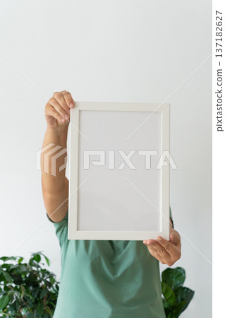 Adult holding blank white frame in green shirt with green plant in background. Modern minimalist design, empty template for text Adult holding blank white frame in green shirt with green plant in background. Modern minimalist design, empty template for text 137182627