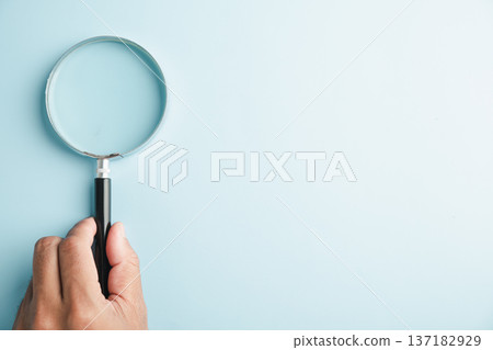 A hand holding a magnifying glass on a light blue background. A hand holding a magnifying glass on a light blue background. 137182929