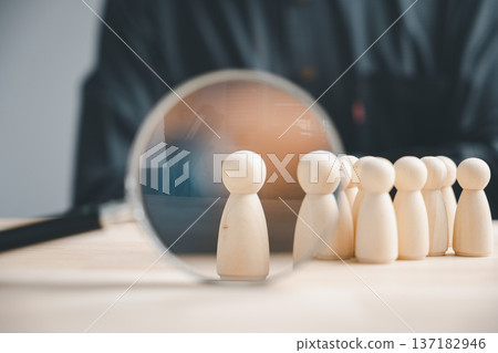 A magnifying glass focuses on a single wooden figurine, standing out from a row of others. 137182946