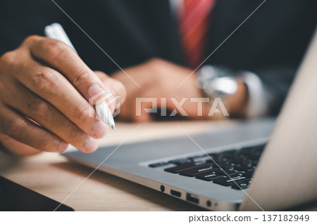 Close-up of a hand holding a white pen over a laptop keyboard. The focus is on the pen. 137182949