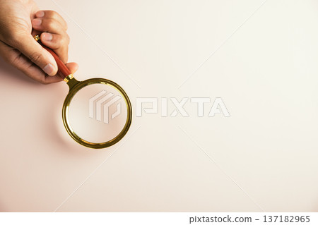 A hand holds a magnifying glass with a gold frame on a light pink background. 137182965