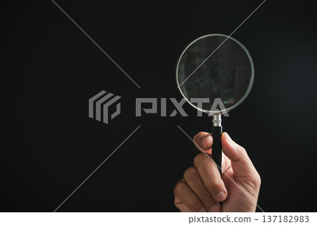 Hand holding a magnifying glass against a black background. 137182983