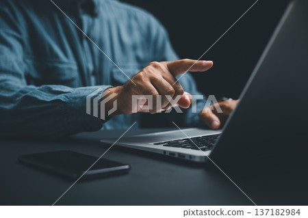 Close-up of a hand pointing at a laptop screen, with a smartphone lying on the table.  Focus on the hand and laptop. 137182984