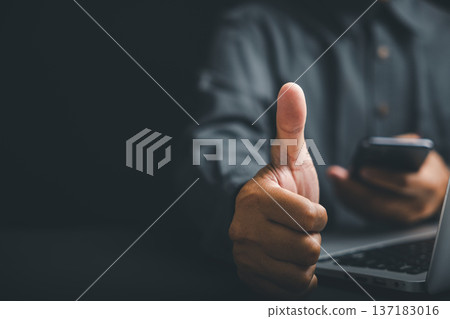 Close-up of a hand giving a thumbs up, holding a smartphone and laptop on a dark background. 137183016