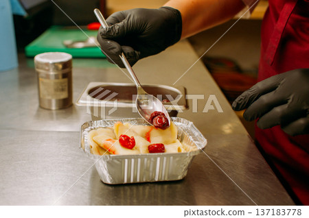 Serving food. Catering kitchen. Chef hand in gloves adding cherries to sweet berry dough meal. Portioned dumpling in takeout foil form. 137183778