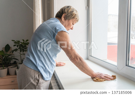 Senior woman performing domestic chores, carefully wiping dust from a window surface during daily routine Senior woman performing domestic chores, carefully wiping dust from a window surface during daily routine 137185006