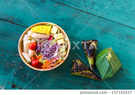 Healthy colorful vegetable salad in a paper bowl and grilled banana leaf wraps on a rustic wooden table. Healthy colorful vegetable salad in a paper bowl and grilled banana leaf wraps on a rustic wooden table. 137185037