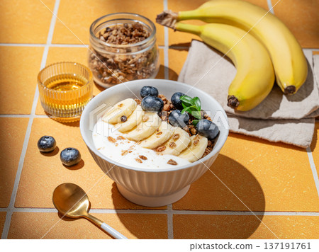 Natural greek yogurt with granola, blueberry and banana in a bowl on a orange tile background Natural greek yogurt with granola, blueberry and banana in a bowl on a orange tile background 137191761