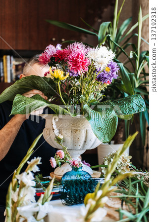 Man arranges colorful aster flowers and large hosta leaves in white urn. Focus-based therapy, mental health for men, hobbyist mastery, cognitive well-being. Man arranges colorful aster flowers and large hosta leaves in white urn. Focus-based therapy, mental health for men, hobbyist mastery, cognitive well-being. 137192318