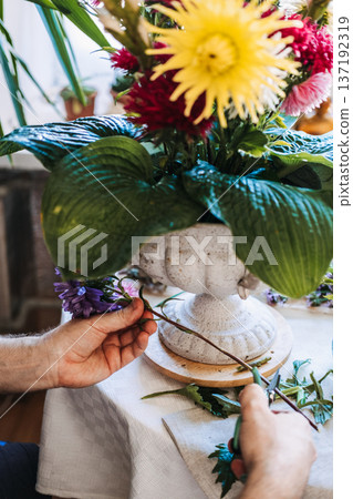 Hands of man trim flower stem with garden pruners next to stone urn. Floral arrangement process, tactile artistry, boutique florist studio, independent creator. 137192319
