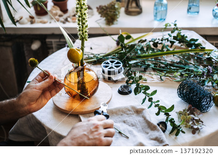 Hands hold yellow fruit on stick near amber glass vase with floral arrangement on table. Contemporary floral styling, botanical art, unique plant arrangements, artistic floristry. Hands hold yellow fruit on stick near amber glass vase with floral arrangement on table. Contemporary floral styling, botanical art, unique plant arrangements, artistic floristry. 137192320