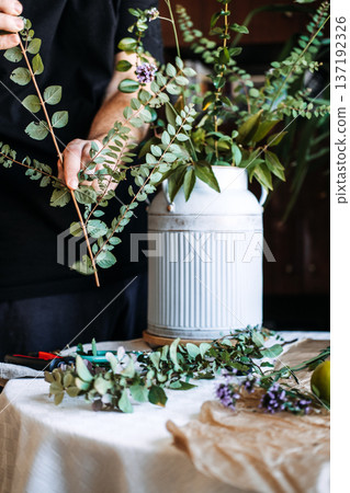 Hands use green garden shears to trim thin botanical stem on table next to white fluted jug. biophilia, nature-inspired living, plant-human connection, green interiors, botanical accents 137192326
