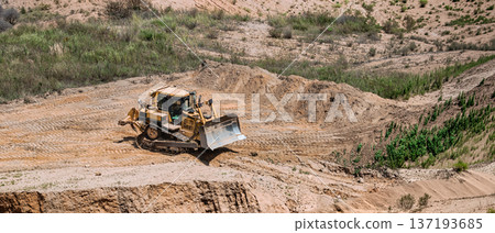 A bulldozer is moving dirt at a construction site in a rural area. The sun is shining over the hills and small plants are growing nearby A bulldozer is moving dirt at a construction site in a rural area. The sun is shining over the hills and small plants are growing nearby 137193685