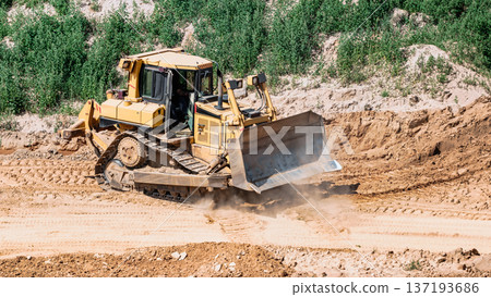 A bulldozer moves dirt at a construction site. Dust swirls as the machine clears the ground in a rural area under a sunny sky A bulldozer moves dirt at a construction site. Dust swirls as the machine clears the ground in a rural area under a sunny sky 137193686