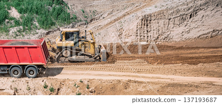A bulldozer is pushing dirt while a dump truck waits nearby at a construction site. Heavy machinery is actively performing tasks in an open area 137193687