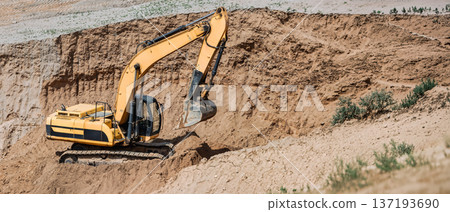 An excavator digs deep into the ground at a construction site, moving dirt and shaping the land under clear blue skies 137193690