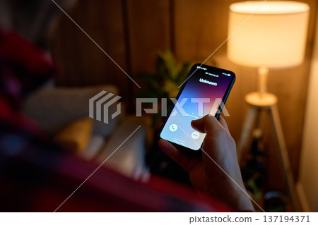 Close-up of hand holding smartphone showing unknown incoming call in living room. Concept of spam call, telemarketing, scam risk and mobile communication Close-up of hand holding smartphone showing unknown incoming call in living room. Concept of spam call, telemarketing, scam risk and mobile communication 137194371