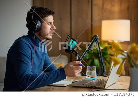 Man wearing headphones recording podcast with microphone and laptop at home desk. Concept of podcasting, online content creation, remote work and audio recording. 137194375