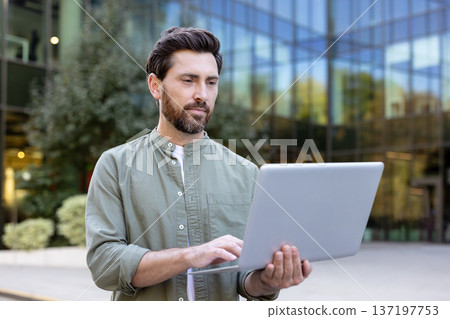 Man standing outdoors in front of a glass building, focused on typing on a laptop, representing remote work, flexible jobs, and mobile business through technology 137197753
