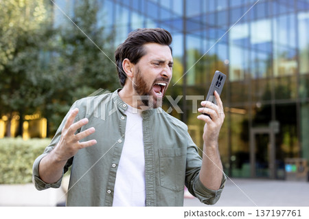 Bearded man shouting at his smartphone outdoors, expressing anger, frustration and stress from digital communication, bad news or a heated argument, intense and upset emotions Bearded man shouting at his smartphone outdoors, expressing anger, frustration and stress from digital communication, bad news or a heated argument, intense and upset emotions 137197761