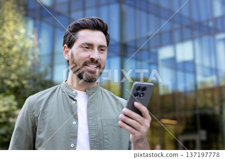 Smiling man in casual wear receiving good news on smartphone, standing against a modern glass building, representing mobile communication and digital connectivity in an urban setting 137197778