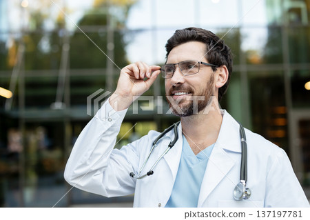 Male doctor in medical uniform wearing eyeglasses and stethoscope, enjoying fresh air while smiling outside a modern hospital building, representing healthcare and professionalism 137197781