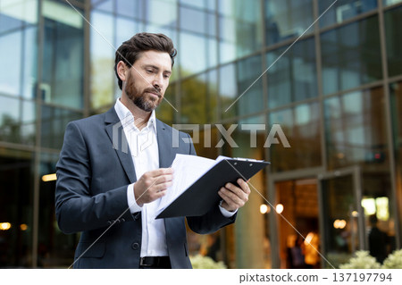 Young professional businessman in a sleek suit carefully reviewing reports and documents on a clipboard while standing outside a contemporary glass corporate building Young professional businessman in a sleek suit carefully reviewing reports and documents on a clipboard while standing outside a contemporary glass corporate building 137197794
