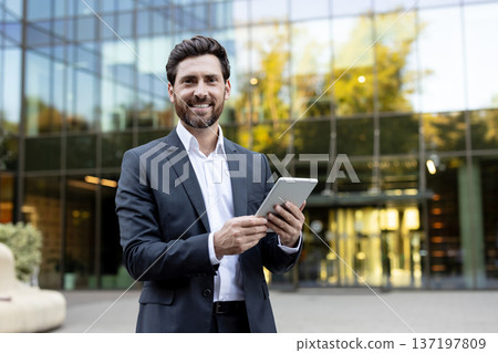 Professional businessman wearing a suit and white shirt smiling at the camera while holding a digital tablet with both hands, standing outdoors in front of a modern glass building Professional businessman wearing a suit and white shirt smiling at the camera while holding a digital tablet with both hands, standing outdoors in front of a modern glass building 137197809