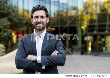 Young professional businessman with a confident smile and arms crossed, standing outdoors in front of a modern corporate office building, symbolizing leadership and success in business 137197834