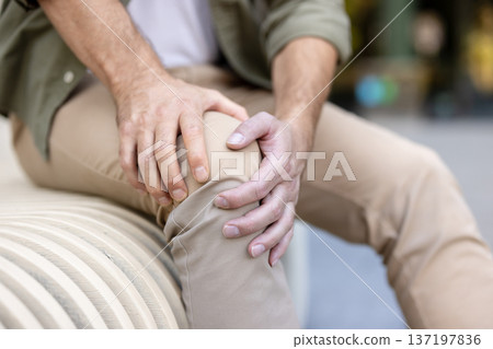 Man crouching outdoors, clutching aching knee in both hands, showing joint pain and discomfort from injury, arthritis or strain, partial close-up, casual clothing, adult male Man crouching outdoors, clutching aching knee in both hands, showing joint pain and discomfort from injury, arthritis or strain, partial close-up, casual clothing, adult male 137197836
