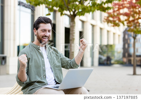 Excited man cheering success while looking at his laptop, sitting on a bench in an urban outdoor setting, personifying achievement, victory, and job well-done 137197885