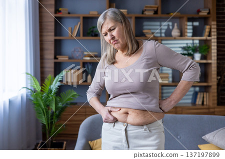 Elderly woman examines stomach with concerned expression. Emphasis on health, wellness, aging, and self-reflection. Scene captures thoughtful awareness of body changes and their emotional impact. Elderly woman examines stomach with concerned expression. Emphasis on health, wellness, aging, and self-reflection. Scene captures thoughtful awareness of body changes and their emotional impact. 137197899
