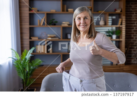 Mature woman celebrating weight loss success with oversized pants in living room. Smiling and giving thumbs-up, expressing joy and confidence. Embracing fitness and healthy lifestyle changes. Mature woman celebrating weight loss success with oversized pants in living room. Smiling and giving thumbs-up, expressing joy and confidence. Embracing fitness and healthy lifestyle changes. 137197917