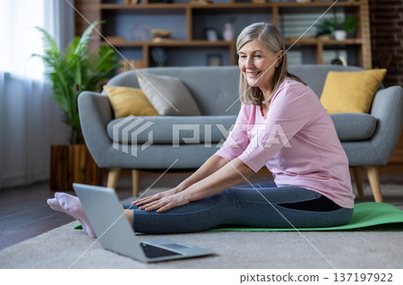 Smiling senior woman practicing yoga at home, using laptop for online class. Focus on health, fitness, flexibility, and relaxation. Peaceful home environment with casual comfort and positivity. Smiling senior woman practicing yoga at home, using laptop for online class. Focus on health, fitness, flexibility, and relaxation. Peaceful home environment with casual comfort and positivity. 137197922
