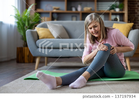 Senior woman with knee pain sits on floor while exercising at home, expressing discomfort and concern. Relaxing in living room, she rests on green yoga mat, wearing casual attire. Senior woman with knee pain sits on floor while exercising at home, expressing discomfort and concern. Relaxing in living room, she rests on green yoga mat, wearing casual attire. 137197934