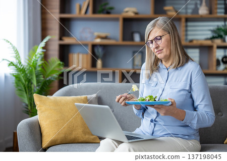 Elderly woman sitting on couch, eating salad, looking dissatisfied, working on laptop at home. Reflects feelings of frustration with healthy diet, multitasking, home office. No background features. Elderly woman sitting on couch, eating salad, looking dissatisfied, working on laptop at home. Reflects feelings of frustration with healthy diet, multitasking, home office. No background features. 137198045